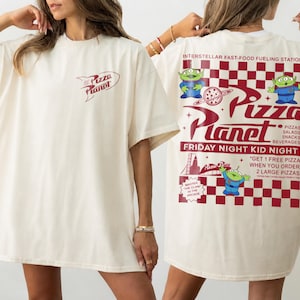 May include: Cream-colored t-shirt with the Pizza Planet logo on the front and back. The back features the text "Interstellar Fast-Food Fueling Station" and "Friday Night Kid Night" with alien characters and pizza graphics.