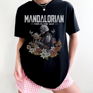 May include: Black t-shirt featuring the text "MANDALORIAN - THIS IS THE WAY" in white. The graphic includes an illustration of the Mandalorian and Grogu surrounded by flowers and butterflies. The shirt is a crew neck.