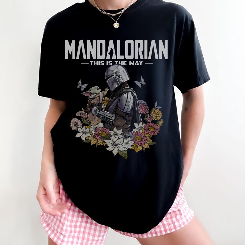 May include: Black t-shirt featuring the text "MANDALORIAN - THIS IS THE WAY" in white. The graphic includes an illustration of the Mandalorian and Grogu surrounded by flowers and butterflies. The shirt is a crew neck.