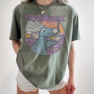 May include: Olive green t-shirt featuring a circular graphic from the movie Ratatouille. The graphic includes the text "La Ratatouille" above a cartoon rat wearing a chef's hat, with the Eiffel Tower in the background. The shirt has short sleeves.