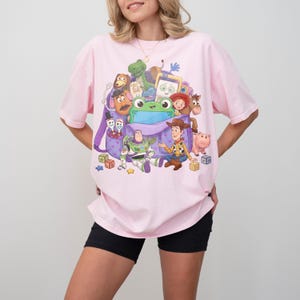 May include: A light pink t-shirt featuring a colorful graphic of characters from the animated movie Toy Story. The graphic includes Woody, Buzz Lightyear, and other characters. The shirt is a relaxed fit.