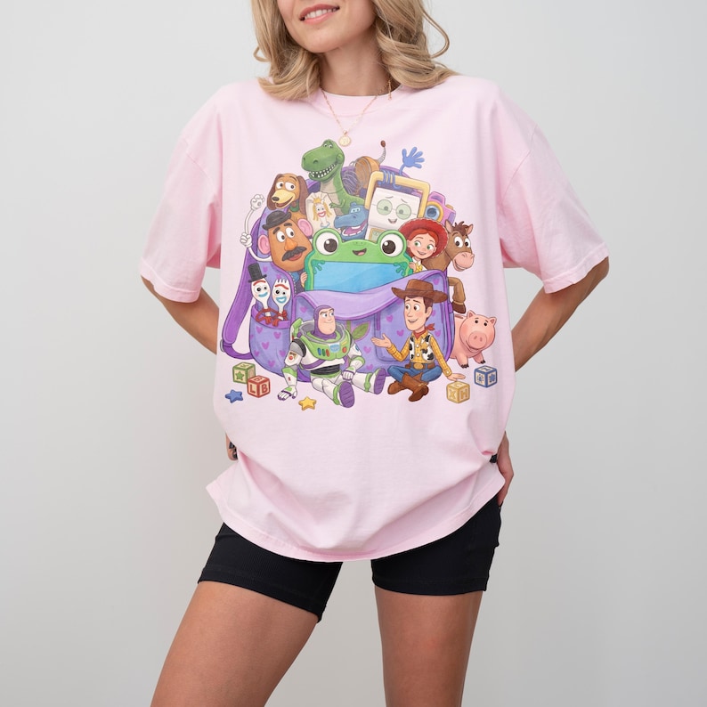 May include: A light pink t-shirt featuring a colorful graphic of characters from the animated movie Toy Story. The graphic includes Woody, Buzz Lightyear, and other characters. The shirt is a relaxed fit.