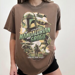 May include: Brown t-shirt featuring a graphic from the Star Wars series, "The Mandalorian and Grogu." The design includes the Mandalorian's helmet, Grogu, and the phrase "THIS IS THE WAY."