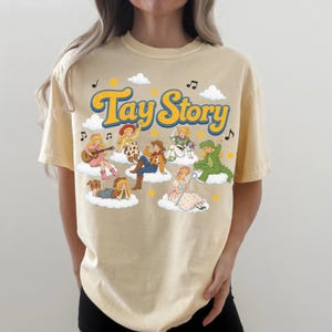 Puede incluir: Una camiseta de manga corta de color beige claro con un estampado gr&aacute;fico que incluye el texto Tay Story en letras azules y amarillas. El dise&ntilde;o muestra varios personajes ilustrados con diversos disfraces sentados sobre nubes blancas con estrellas.