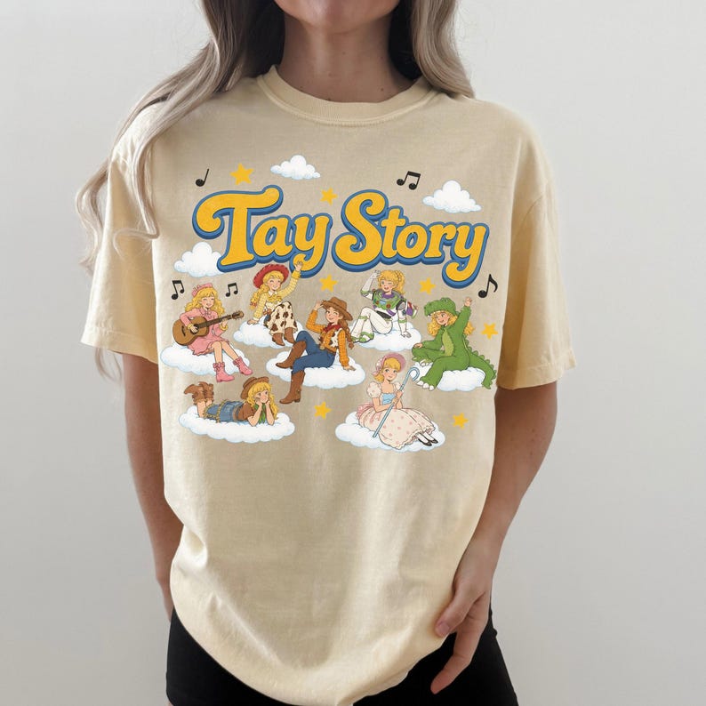 Puede incluir: Una camiseta de manga corta de color beige claro con un estampado gr&aacute;fico que incluye el texto Tay Story en letras azules y amarillas. El dise&ntilde;o muestra varios personajes ilustrados con diversos disfraces sentados sobre nubes blancas con estrellas.