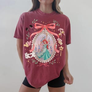 Pu&ograve; includere: T-shirt bordeaux con un'illustrazione incorniciata di Ariel de La Sirenetta in un abito turchese, con un castello sullo sfondo. Il design include un fiocco rosa, orecchie di Topolino e altri elementi decorativi. La maglietta ha una vestibilit&agrave; ampia.