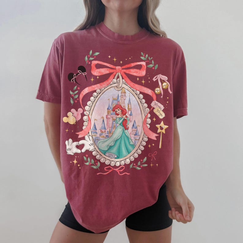 Pu&ograve; includere: T-shirt bordeaux con un'illustrazione incorniciata di Ariel de La Sirenetta in un abito turchese, con un castello sullo sfondo. Il design include un fiocco rosa, orecchie di Topolino e altri elementi decorativi. La maglietta ha una vestibilit&agrave; ampia.