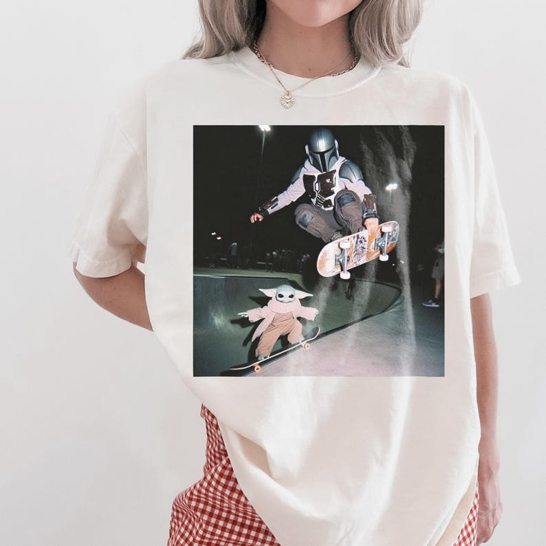 Pu&ograve; includere: Una t-shirt color bianco sporco con una stampa grafica di un personaggio con casco e armatura argentati che esegue un trick con lo skateboard in aria, mentre una piccola creatura verde con una tunica rosa va sullo skateboard in uno skatepark.