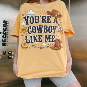 May include: A yellow short-sleeve t-shirt featuring the dark blue text "YOU'RE A COWBOY LIKE ME" surrounded by illustrations of a brown cowboy hat, a sheriff badge, a rocket ship, a colorful ball, clouds, and stars in various colors.