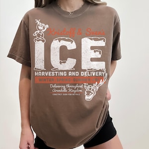 May include: Brown t-shirt featuring a distressed graphic with the text "Kristoff & Sven's ICE Harvesting and Delivery." The design includes images of Olaf and Sven from the movie Frozen. The shirt also has the text "Delivering throughout Arendelle Kingdom."