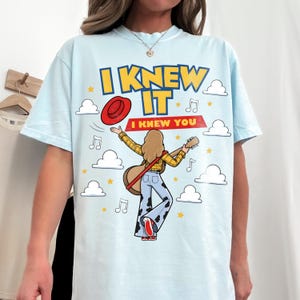 May include: A light blue t-shirt featuring a graphic of a person with long hair playing a guitar, wearing a yellow plaid shirt and cow-print bell-bottom pants. The text reads I KNEW IT and I KNEW YOU, surrounded by clouds, stars, and music notes.