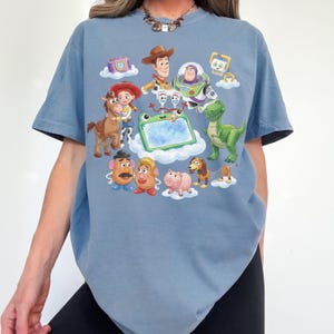 May include: A light blue t-shirt featuring a colorful graphic of characters from the animated movie Toy Story. The design includes Woody, Buzz Lightyear, Jessie, Rex, Mr. Potato Head, and others. The shirt has a crew neck and short sleeves.