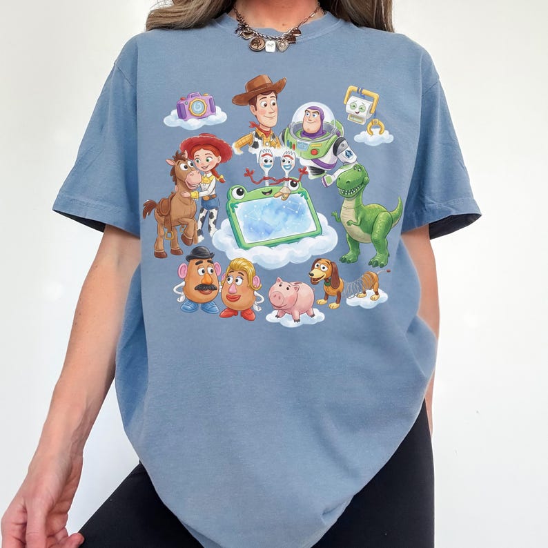 May include: A light blue t-shirt featuring a colorful graphic of characters from the animated movie Toy Story. The design includes Woody, Buzz Lightyear, Jessie, Rex, Mr. Potato Head, and others. The shirt has a crew neck and short sleeves.
