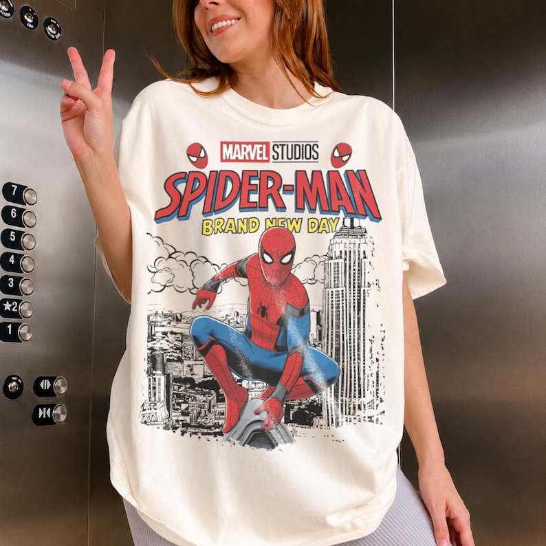 May include: An off-white t-shirt featuring a graphic print of Spider-Man in a red and blue suit crouching over a city skyline. The text reads Marvel Studios Spider-Man Brand New Day, with small Spider-Man mask icons above the title text.