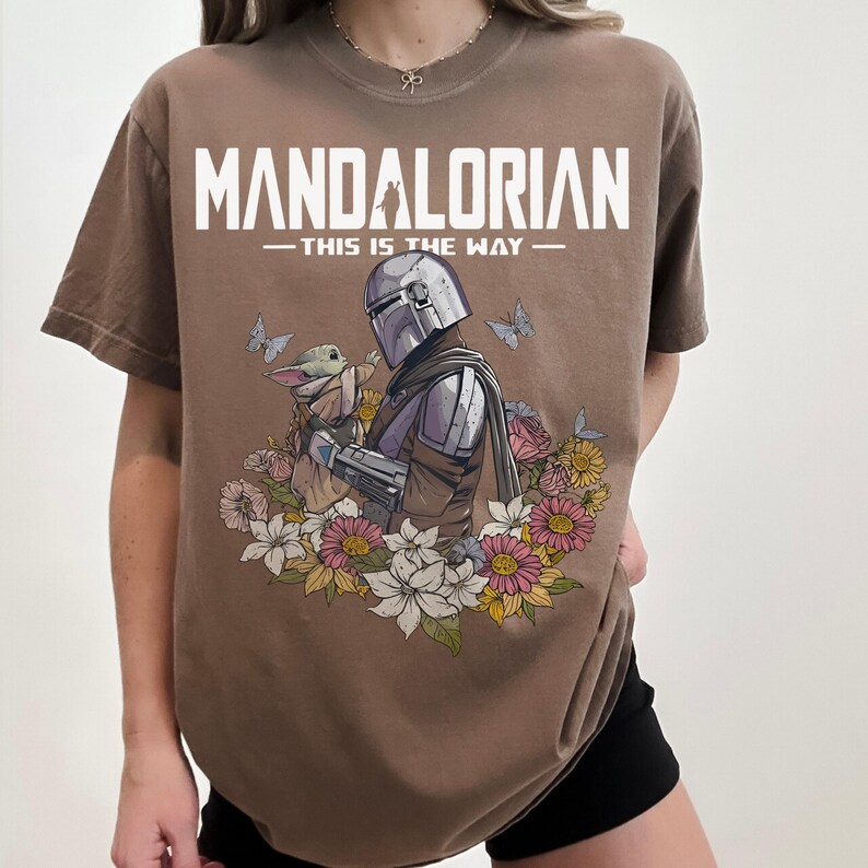 May include: Brown t-shirt featuring a graphic of The Mandalorian and Grogu surrounded by flowers and butterflies. The text "MANDALORIAN - THIS IS THE WAY" is printed across the top in white. The shirt is a crew neck.