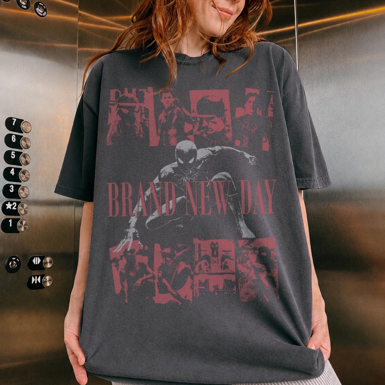 May include: A charcoal grey short-sleeve t-shirt featuring a large graphic print in faded red. The design includes a central image of a superhero in a crouched pose, surrounded by smaller panels of figures and the text BRAND NEW DAY in bold letters.