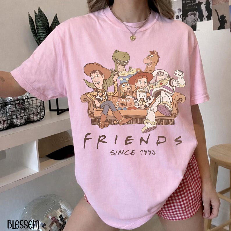 May include: A pale pink t-shirt with a graphic of Toy Story characters on a sofa. The word "FRIENDS" is printed below the graphic, with "SINCE 1995" underneath. The shirt is worn with red and white checked shorts.