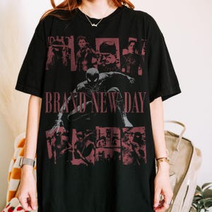 May include: A black short-sleeved t-shirt featuring a large graphic print of a superhero in a crouching pose. The text BRAND NEW DAY is printed in red across the centre, surrounded by multiple smaller red-toned panels depicting various scenes.