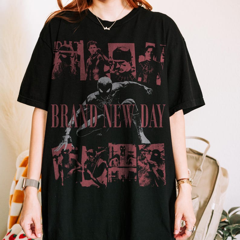 May include: A black short-sleeved t-shirt featuring a large graphic print of a superhero in a crouching pose. The text BRAND NEW DAY is printed in red across the centre, surrounded by multiple smaller red-toned panels depicting various scenes.