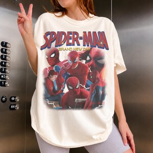May include: An off-white short-sleeve t-shirt featuring a graphic print of Spider-Man in various poses. The text "SPIDER-MAN" appears in large blue and red letters at the top, with "BRAND NEW DAY" printed underneath in smaller yellow font.