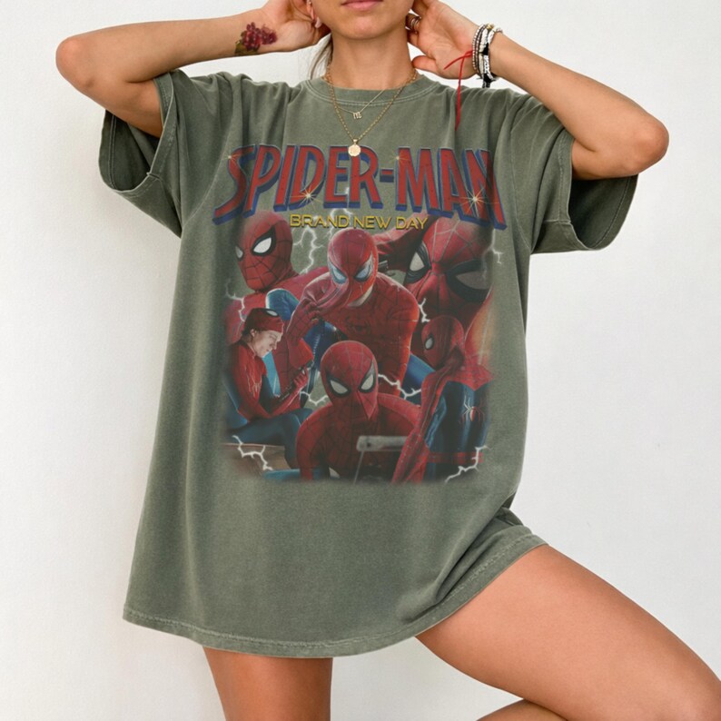 May include: An olive green short-sleeve t-shirt featuring a large graphic print of the character Spider-Man in various poses. The text "SPIDER-MAN" appears in bold red letters at the top, with the words "BRAND NEW DAY" printed in yellow below.