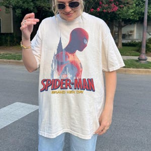 May include: A cream-colored short-sleeve t-shirt featuring a graphic of Spider-Man in profile against a city skyline. The text reads Marvel Studios Spider-Man Brand New Day in red and yellow lettering across the center of the chest area.