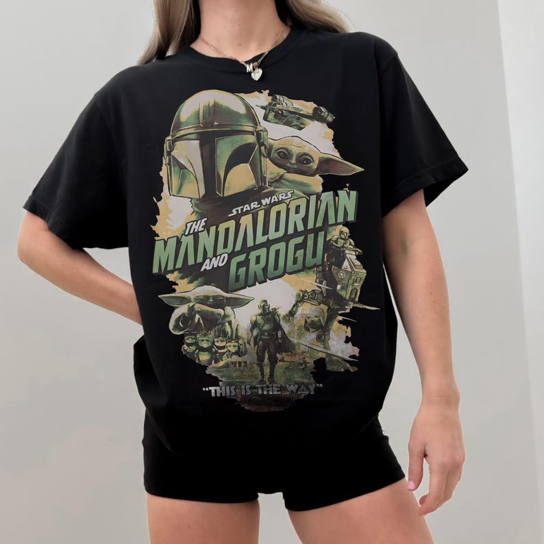 May include: Black t-shirt featuring a graphic from the Star Wars series, "The Mandalorian and Grogu." The design includes the Mandalorian's helmet, Grogu, and the phrase "This is the Way."