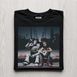 以下が含まれることがあります： 黒いTシャツ。トイ・ストーリーのバズ・ライトイヤーとウッディが縁石に座っているグラフィック。バズはボトルから飲んでいます。「Comfort」の文字が襟にプリントされています。