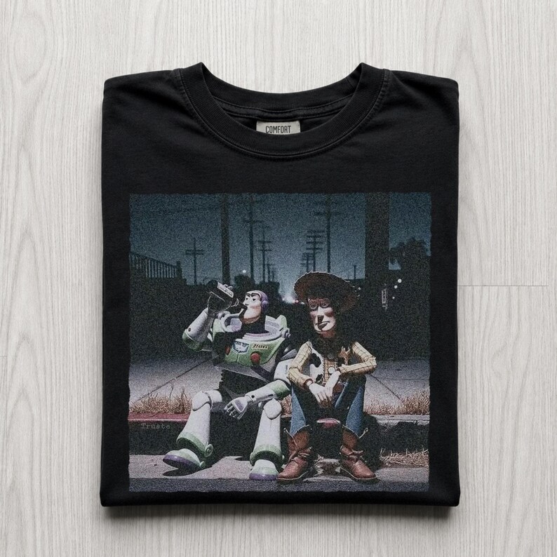 以下が含まれることがあります： 黒いTシャツ。トイ・ストーリーのバズ・ライトイヤーとウッディが縁石に座っているグラフィック。バズはボトルから飲んでいます。「Comfort」の文字が襟にプリントされています。