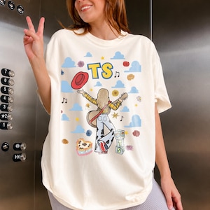 May include: An off-white t-shirt featuring a graphic print of a person with blonde hair wearing a yellow plaid shirt and cow-print trousers, holding a guitar and a red hat. The design includes blue clouds, musical notes, flowers, and blocks with the text TS.