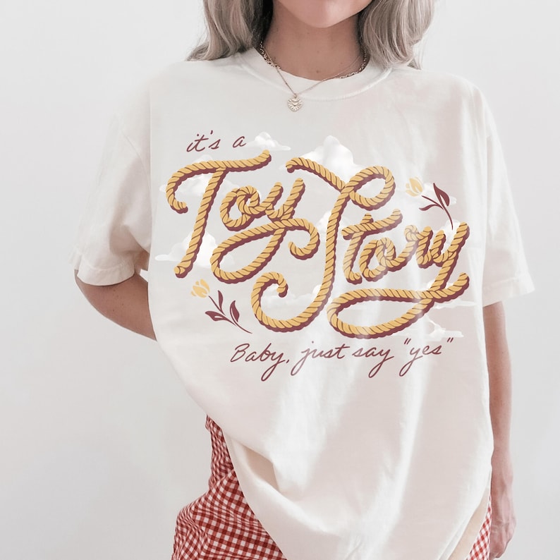 May include: An off-white short-sleeve t-shirt featuring a graphic design with the text "it's a Toy Story" written in a rope-like font, accompanied by the phrase "Baby, just say 'yes'" in a dark red script, with small floral and cloud accents.
