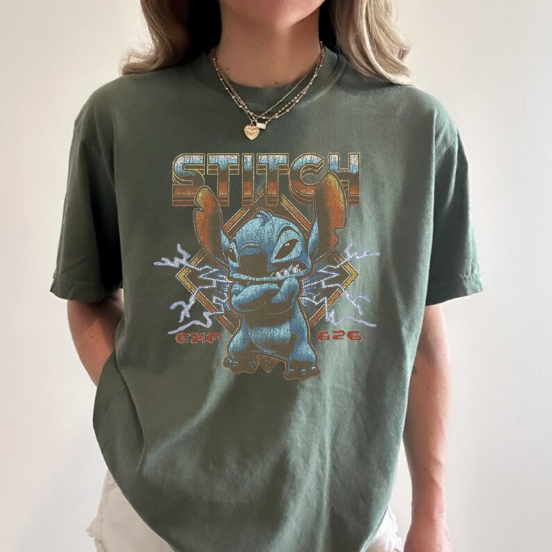 May include: Olive green t-shirt featuring a graphic of the character Stitch with the word "STITCH" above him. The graphic includes lightning bolts and the numbers "626" and "626" on either side of Stitch.