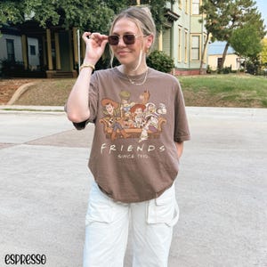 May include: Espresso-coloured t-shirt with a graphic of Toy Story characters on a sofa, with the word "FRIENDS" and "SINCE 1995" printed below. The model is wearing white trousers and sunglasses.
