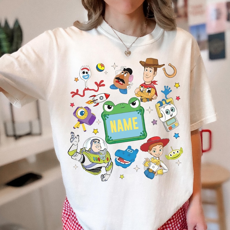 May include: A cream-colored t-shirt featuring a colorful graphic print of various animated toy characters, including a cowboy, a space ranger, a cowgirl, a potato figure, and a fork character, surrounding a central green frog-shaped frame with the text NAME.