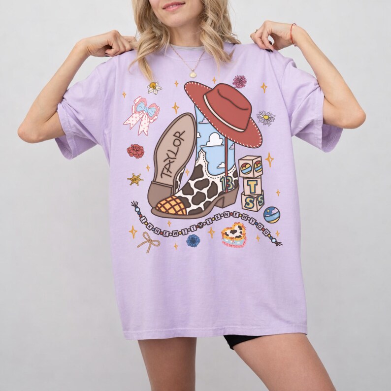 May include: A light purple t-shirt featuring a graphic print of a cowboy boot with a cow print pattern, a red cowboy hat, a stack of blocks with the letters T, S, and the number 13, a beaded bracelet, and various small floral and star icons.
