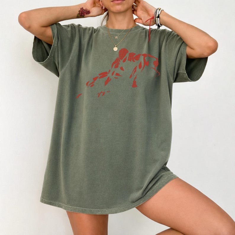 May include: An olive green short-sleeved t-shirt featuring a red graphic print of a crouching figure on the chest. The garment has a crew neckline and a relaxed, loose-fitting silhouette, constructed from a solid-colored cotton fabric.