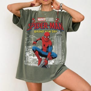 May include: An olive green short-sleeve t-shirt featuring a graphic print of Spider-Man in a crouching pose. The design includes the text Marvel Studios Spider-Man Brand New Day above a black and white illustration of a city skyline.
