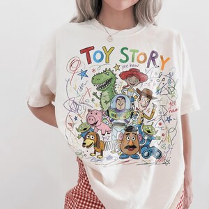 May include: Cream-colored t-shirt featuring a colorful graphic of Toy Story characters. The design includes the words "TOY STORY" in rainbow colors, along with drawings of Buzz Lightyear, Woody, and other characters. The shirt has a crew neck and short sleeves.