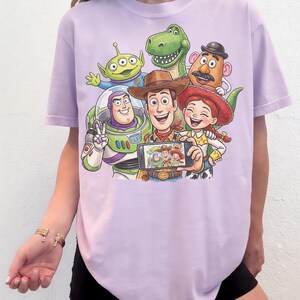 May include: A light purple t-shirt featuring a graphic of characters from the animated movie Toy Story. The characters include Woody, Buzz Lightyear, Jessie, Rex, Mr. Potato Head, and an alien, all posing for a selfie.