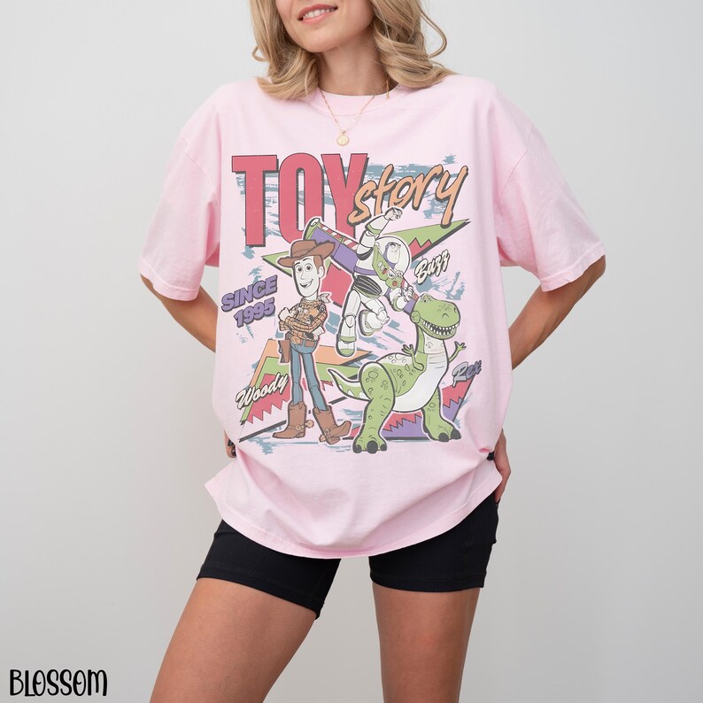 Pode incluir: T-shirt rosa claro com uma estampa gr&aacute;fica de personagens do filme "Toy Story". O design inclui Woody, Buzz Lightyear e Rex, com as palavras "Toy Story" e "Since 1995". A camiseta &eacute; de manga curta e com corte folgado.