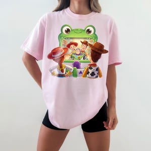 Puede incluir: Camiseta rosa claro con un dise&ntilde;o de personajes de la pel&iacute;cula de animaci&oacute;n Toy Story. El dise&ntilde;o muestra a Woody, Buzz Lightyear, Jessie y otros personajes en una m&aacute;quina recreativa verde con forma de rana.
