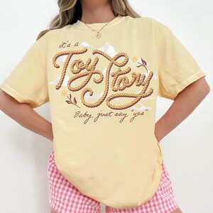 May include: A pale yellow short-sleeve t-shirt featuring a graphic design with the text "it's a Toy Story" written in a rope-like font, accompanied by the phrase "Baby, just say "yes"" and small floral illustrations against a cloud background.