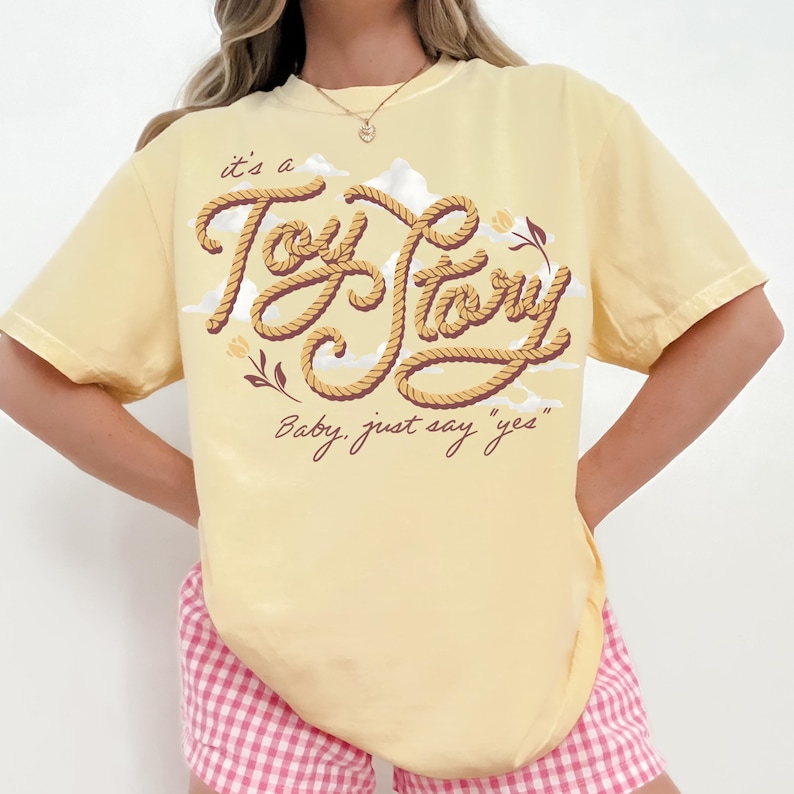 May include: A pale yellow short-sleeve t-shirt featuring a graphic design with the text "it's a Toy Story" written in a rope-like font, accompanied by the phrase "Baby, just say "yes"" and small floral illustrations against a cloud background.