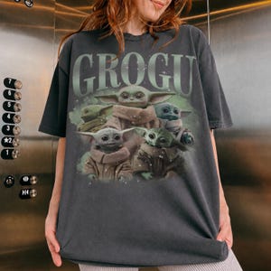 May include: Dark gray t-shirt featuring a graphic of Grogu, also known as Baby Yoda, surrounded by other Grogu characters. The word "GROGU" is printed in a green, stylized font above the image. The shirt is a short-sleeved, crew neck style.