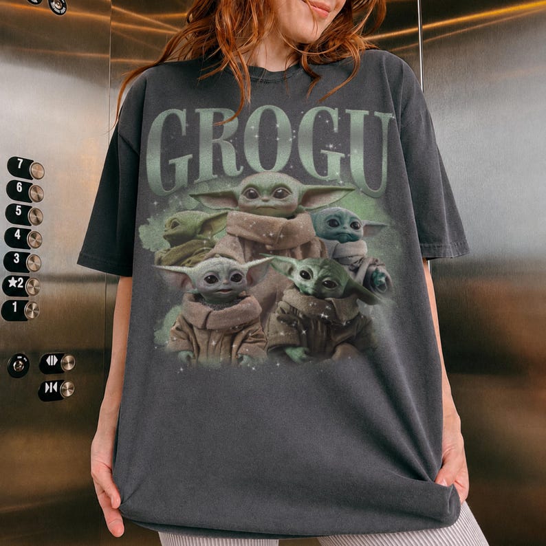 May include: Dark gray t-shirt featuring a graphic of Grogu, also known as Baby Yoda, surrounded by other Grogu characters. The word "GROGU" is printed in a green, stylized font above the image. The shirt is a short-sleeved, crew neck style.