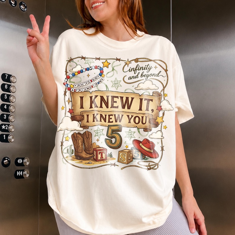 May include: An off-white t-shirt featuring a graphic print with a brown scroll banner that reads "I KNEW IT, I KNEW YOU" above the number "5". The design includes cowboy boots, a red hat, alphabet blocks, a beaded bracelet, and "Cinfinity and beyond".