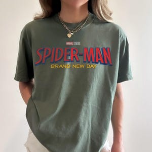 May include: An olive green short-sleeve t-shirt featuring the Marvel Studios logo above the red and blue text "SPIDER-MAN" and the yellow text "BRAND NEW DAY" printed across the chest. The shirt has a crew neckline and a relaxed fit.