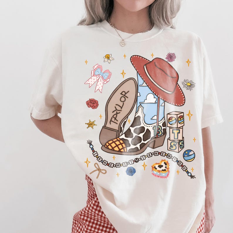 May include: An off-white t-shirt featuring a graphic print of a cowboy boot with a cow print pattern, a red hat, blocks spelling T S, and a beaded string with the text I KNEW IT Y I KNEW YOU. The boot sole is labeled TAYLOR.