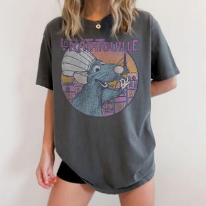 May include: Dark grey t-shirt featuring a circular graphic of a cartoon rat wearing a chef's hat, with the Eiffel Tower in the background. The text "La Ratatouille" is arched above the graphic. The shirt is a relaxed fit.