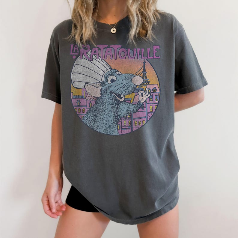May include: Dark grey t-shirt featuring a circular graphic of a cartoon rat wearing a chef's hat, with the Eiffel Tower in the background. The text "La Ratatouille" is arched above the graphic. The shirt is a relaxed fit.